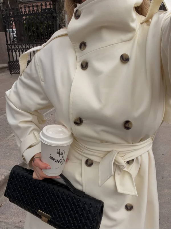 Classic cream colored long trench coat featuring adjustable waist belt and button front